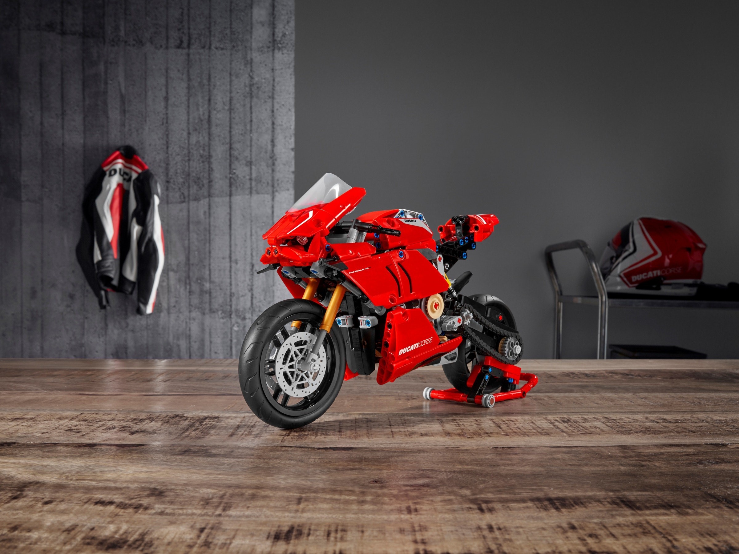 LEGO Technic Ducati Panigale V4 R (42107) on its paddock stand on a wooden table, left front three-quarter view, with a Ducati jacket and helmet in the background