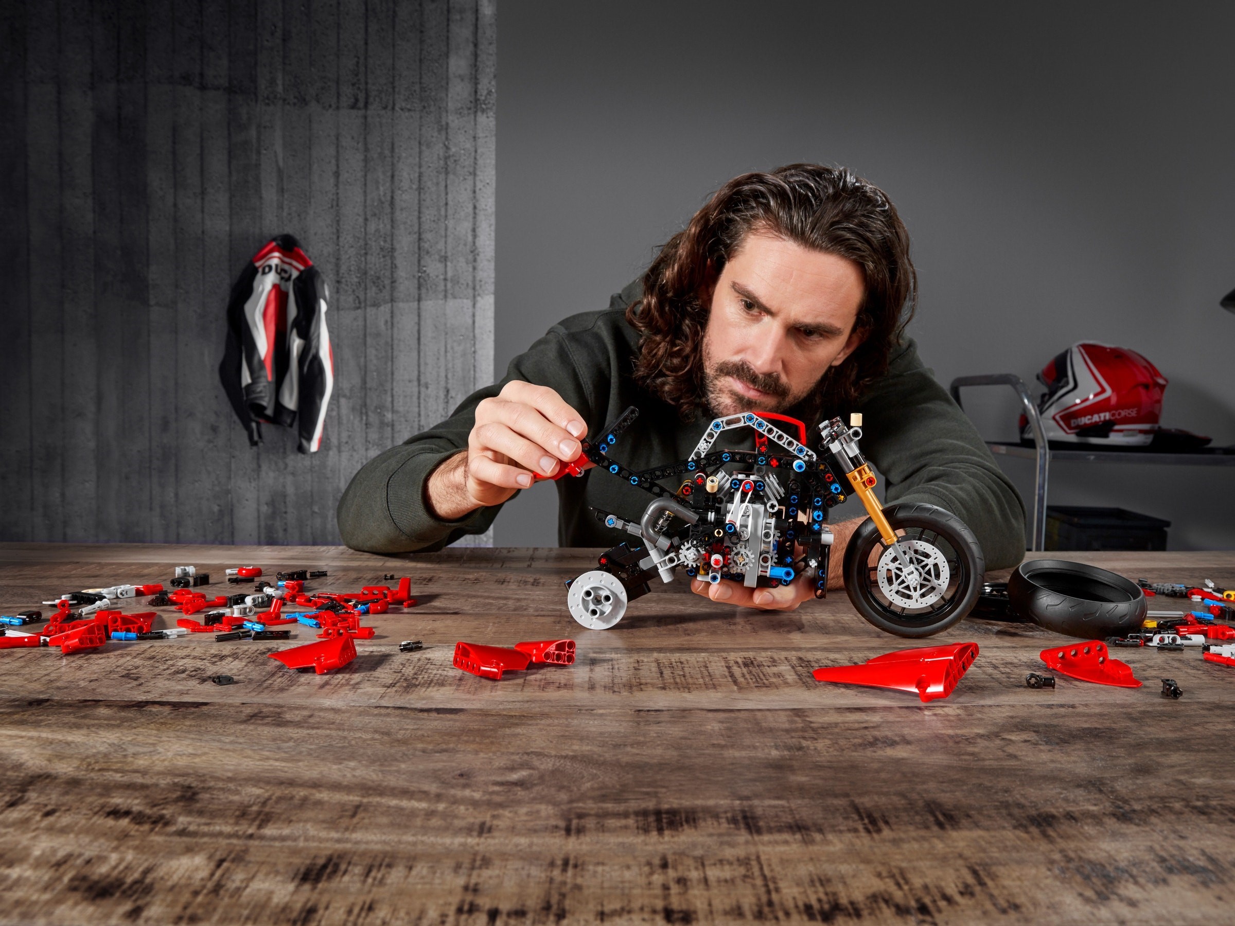 A builder holding the bare chassis of LEGO Technic Ducati Panigale V4 R (42107) part-built: the black frame with the engine and gearbox inside, a gold fork leg and the front wheel fitted, the rear tyre and red panels loose on the table